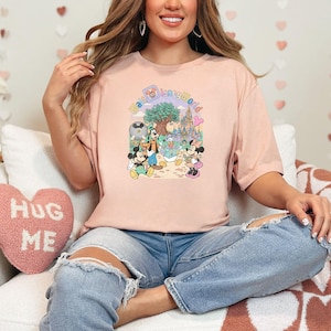 Puede incluir: Una camiseta rosa claro con un gr&aacute;fico colorido de personajes de Disney y el texto "magical world". El dise&ntilde;o incluye Mickey Mouse, Minnie Mouse, Donald Duck y Goofy. La camiseta se usa con jeans azules.