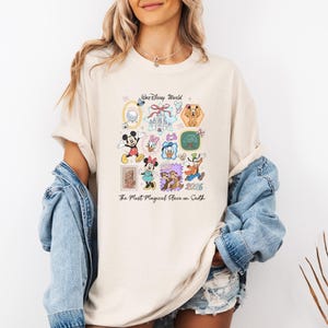 Peut inclure: T-shirt cr&egrave;me avec un motif color&eacute; de personnages et d'ic&ocirc;nes Disney. Le design comprend Mickey Mouse, Minnie Mouse, Donald Duck et Dingo, avec le texte "Walt Disney World" et "The Most Magical Place on Earth 2026."