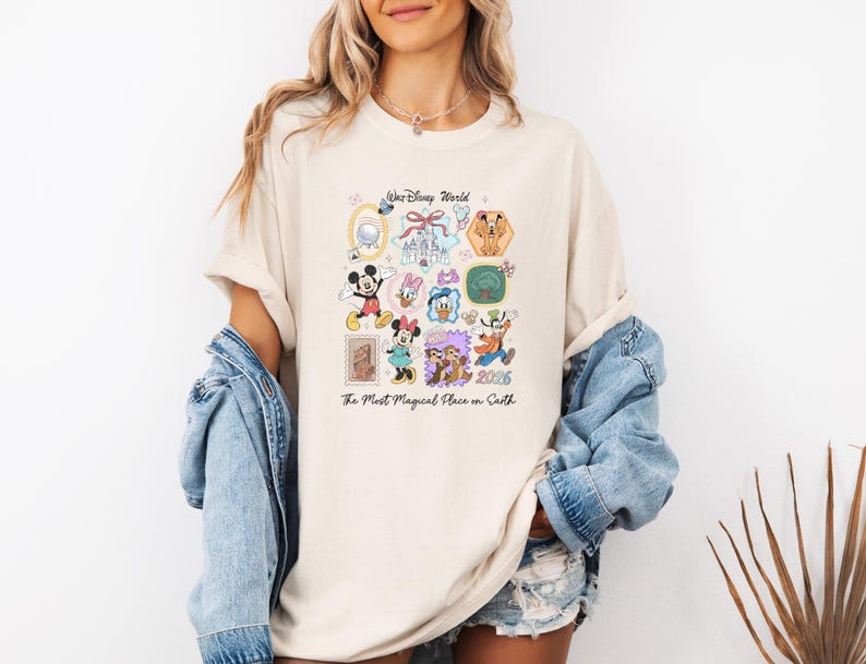 Peut inclure: T-shirt cr&egrave;me avec un motif color&eacute; de personnages et d'ic&ocirc;nes Disney. Le design comprend Mickey Mouse, Minnie Mouse, Donald Duck et Dingo, avec le texte "Walt Disney World" et "The Most Magical Place on Earth 2026."