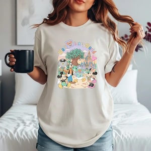 Puede incluir: Camiseta beige con un gr&aacute;fico colorido de personajes de Disney, incluyendo Mickey Mouse, Minnie Mouse, Donald Duck y Goofy, con el texto "Walt Disney World" en un arco iris pastel.