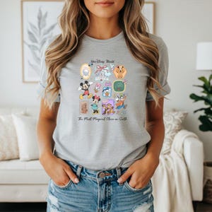 Peut inclure: T-shirt gris clair avec un motif color&eacute; de personnages et d'ic&ocirc;nes de Disney World. Le motif comprend Mickey Mouse, Minnie Mouse, Donald Duck et Dingo, ainsi que le texte "Walt Disney World" et "The Most Magical Place on Earth."