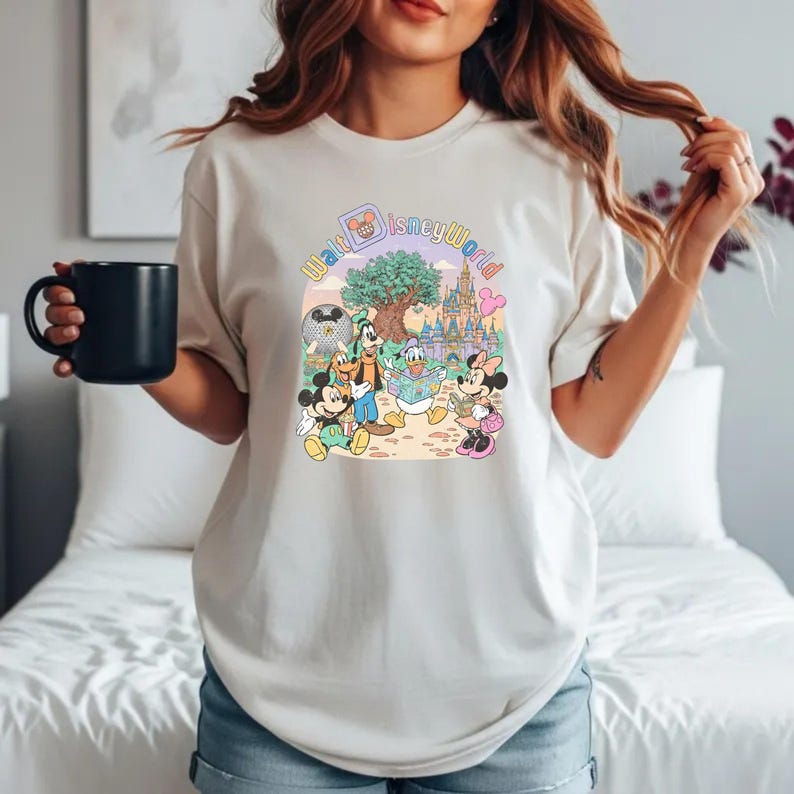 Puede incluir: Camiseta color crema con un gr&aacute;fico colorido de Mickey Mouse, Minnie Mouse, Goofy y Donald Duck frente a un castillo y un &aacute;rbol, con las palabras "Walt Disney World" en un arco&iacute;ris.