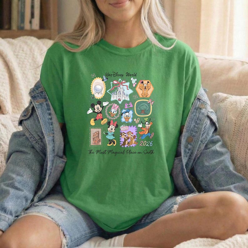 Peut inclure: T-shirt vert avec des personnages et ic&ocirc;nes Disney, dont Mickey Mouse, Donald Duck et le ch&acirc;teau de Cendrillon. Le t-shirt affiche le texte "Walt Disney World" et "2026" avec la phrase "The Most Magical Place on Earth".