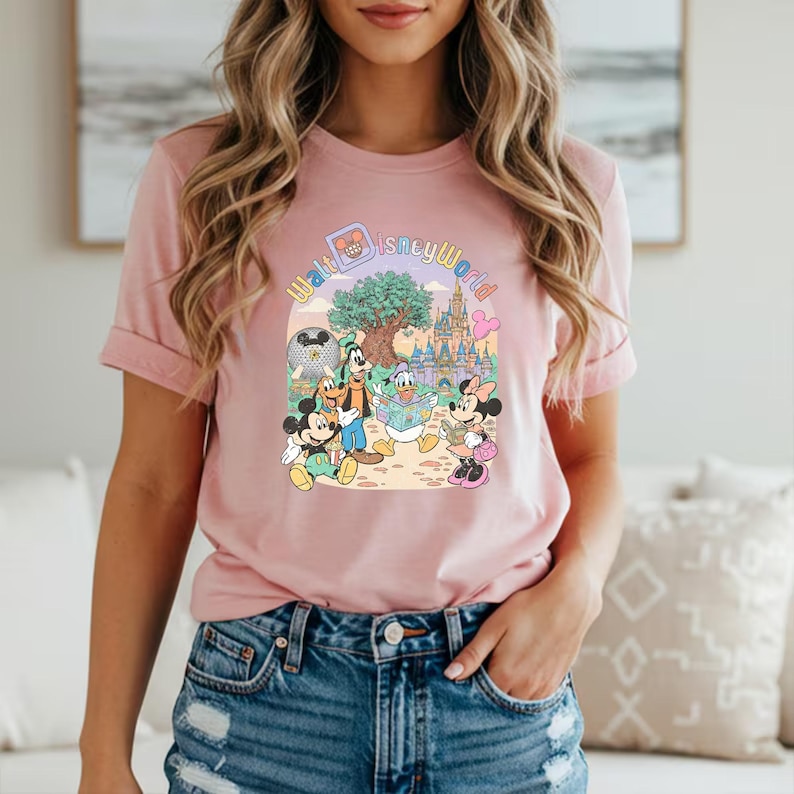 Puede incluir: Camiseta rosa claro con un gr&aacute;fico colorido de personajes de Disney, incluyendo Mickey y Minnie Mouse, Donald Duck y Goofy, con el texto "Walt Disney World" en un arco. El dise&ntilde;o incluye un castillo y un &aacute;rbol.