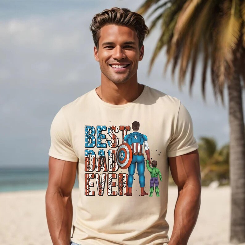 May include: Beige t-shirt with the text "BEST DAD EVER" in blue, red, and white. The graphic depicts a superhero and a child. The superhero is holding the child's hand. The background is a beach scene.