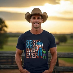 May include: A navy blue t-shirt with the text "BEST DAD EVER" in red, white, and blue. The shirt features a graphic of Captain America and the Hulk. A man wearing a cowboy hat smiles in front of a wooden fence.