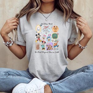 Peut inclure: T-shirt gris clair avec un motif graphique color&eacute; repr&eacute;sentant divers personnages et ic&ocirc;nes Disney. Le texte "Walt Disney World" et "The Most Magical Place on Earth" sont &eacute;galement visibles. Le t-shirt est en mati&egrave;re douce.