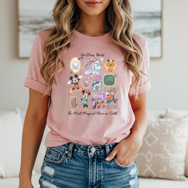 Peut inclure: Un t-shirt rose clair orn&eacute; d'un motif color&eacute; repr&eacute;sentant divers personnages et ic&ocirc;nes de Disney. Le design comprend Mickey Mouse, Minnie Mouse, Donald Duck et Dingo, ainsi que le texte "Walt Disney World" et "The Most Magical Place on Earth 2026".