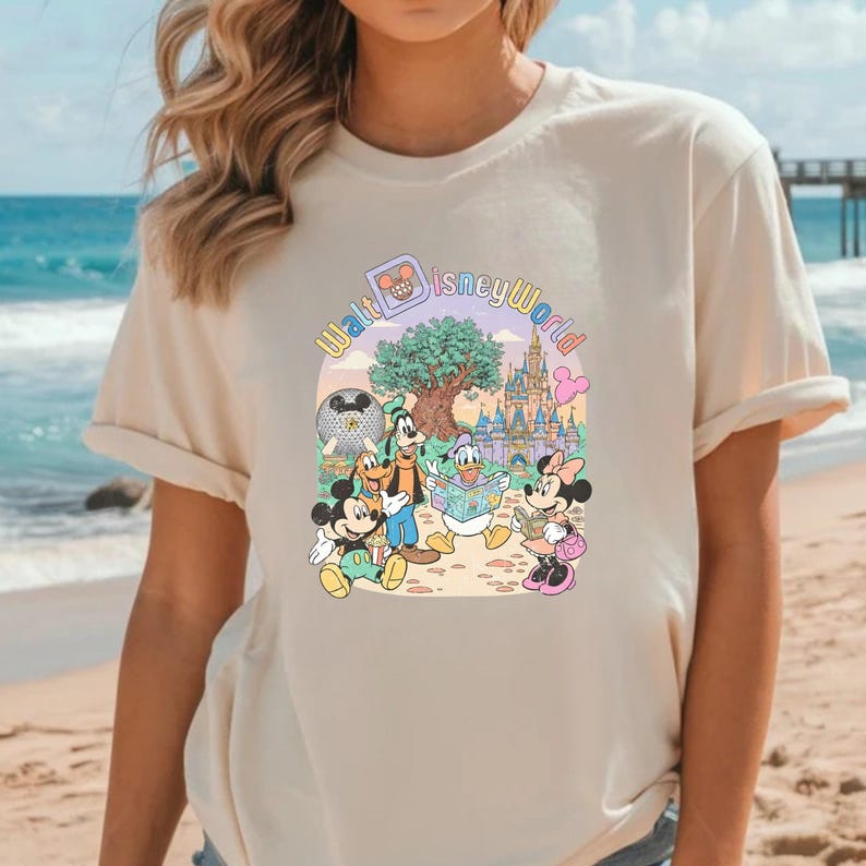 Puede incluir: Camiseta beige con un gr&aacute;fico colorido de personajes de Disney, incluyendo Mickey y Minnie Mouse, Goofy y el Pato Donald, con el texto "Walt Disney World". El dise&ntilde;o incluye un castillo, un &aacute;rbol y un mapa.