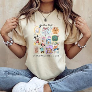 Peut inclure: T-shirt cr&egrave;me avec un motif color&eacute; de Disney World. Le design comprend divers personnages Disney comme Mickey Mouse, Minnie Mouse, Donald Duck et Dingo, ainsi que le texte "Walt Disney World" et "The Most Magical Place on Earth."