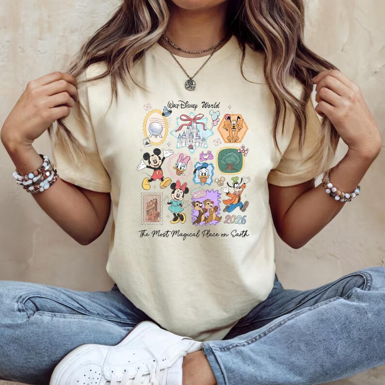 Peut inclure: T-shirt cr&egrave;me avec un motif color&eacute; de Disney World. Le design comprend divers personnages Disney comme Mickey Mouse, Minnie Mouse, Donald Duck et Dingo, ainsi que le texte "Walt Disney World" et "The Most Magical Place on Earth."