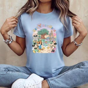 Puede incluir: Camiseta azul claro con un gr&aacute;fico colorido de personajes de Disney, incluyendo Mickey Mouse, Goofy y Minnie Mouse, con el texto "Walt Disney World". El dise&ntilde;o incluye un castillo y un &aacute;rbol.