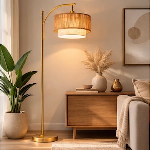 May include: A gold-toned floor lamp with a woven lampshade. The lamp has a curved arm and a round base. The lampshade is cylindrical and has a layered design. A potted plant and a wooden cabinet are in the background.
