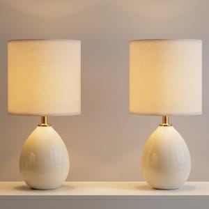 May include: Two matching table lamps. Each lamp has a cream-colored, teardrop-shaped base and a cylindrical lampshade. The lampshades are a light beige color. The lamps have a gold-colored metal accent at the base of the lampshade.