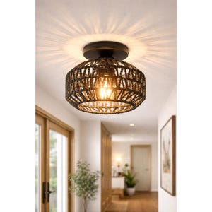 May include: A black woven ceiling light fixture with a warm, glowing bulb. The light is mounted on a dark circular base and casts a patterned light on the ceiling. The fixture is in a hallway with wooden doors and a framed picture.