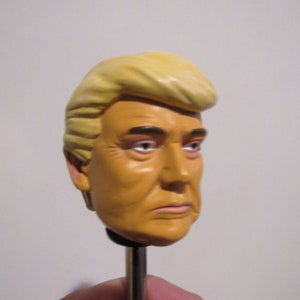 Op de afbeelding: Een geel plastic buste van een persoon met blond haar. De buste staat op een metalen standaard.