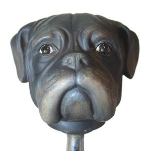 May include: A black and brown bulldog head sculpture, with a realistic look, is mounted on a metal rod. The sculpture is made of a hard material and has a detailed face with a slightly sad expression.