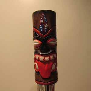 May include: A wooden tiki-style beer tap handle with a carved face, red tongue, and white teeth. The handle is painted with red, white, and blue accents.