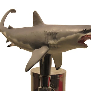 May include: A gray and white shark shaped beer tap handle with an open mouth and sharp teeth. The tap handle is made of plastic and is mounted on a silver metal base.