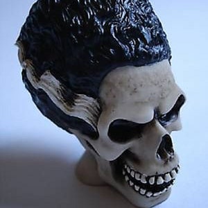 May include: A white skull with black hair sculpted in a Frankenstein style. The skull has a detailed face with teeth and a furrowed brow.