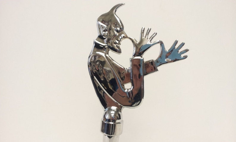 May include: A chrome-plated figurine of a devil with horns and a pointed tail. The devil is holding its hands up in a gesture of surrender or defense. The figurine is a decorative piece that could be used as a bottle stopper or a paperweight.
