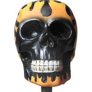 May include: A black and orange skull-shaped bottle stopper. The skull has a black face and a yellow and orange flame design on the top. The teeth are white. The stopper is attached to a dark brown stem.