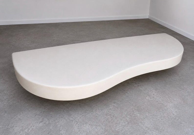 K&ouml;nnte beinhalten: Eine flache, nierenf&ouml;rmige Bank mit einer glatten, matten cremewei&szlig;en Oberfl&auml;che. Das M&ouml;belst&uuml;ck zeichnet sich durch eine durchgehend geschwungene Silhouette mit abgerundeten Kanten und eine solide, nahtlose Konstruktion aus, die bodennah ist.