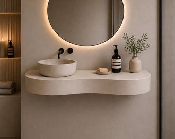 May include: A wall-mounted, curved beige vanity featuring a matching stone vessel sink, a matte black faucet, and a round backlit mirror. The countertop holds a soap dispenser, a small dish with a bar of soap, and a vase with green foliage.