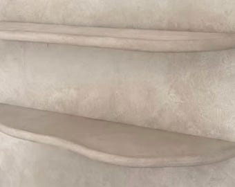 May include: Two wall-mounted floating shelves with a smooth, light beige, matte finish. Each shelf features a curved, organic front edge and a flat top surface, designed to be attached directly to a wall for horizontal storage and display.