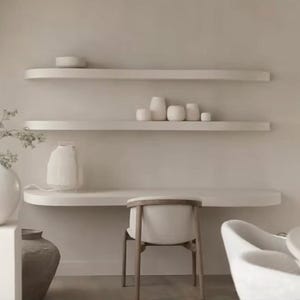 May include: A wall-mounted desk with two matching floating shelves above, all in a matte off-white finish. A wooden chair with a curved backrest sits at the desk. Several small, rounded white ceramic vases are arranged on the shelves.