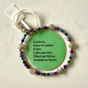 Könnte beinhalten: Ein Armband mit Edelsteinperlen, darunter blauer Kyanit, Rubin und Labradorit. Das Armband ist an einer Silberkette befestigt und hat einen Edelstahlverschluss. Ein grünes Etikett listet die Materialien auf.