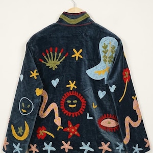 May include: Navy blue velvet jacket with colorful embroidered designs. The jacket features a high collar and is adorned with embroidered stars, suns, boots, and other whimsical shapes. The jacket is hanging on a wooden hanger.