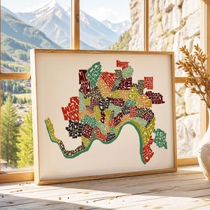 May include: A typography art print of a city map with the names of neighborhoods in different colors and fonts. The map is framed in a light wood frame and is hanging on a wall.