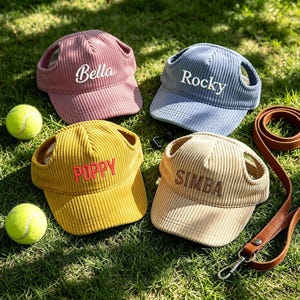 May include: Four corduroy dog hats in pink, blue, yellow, and beige, each embroidered with a name: Bella, Rocky, Poppy, and Simba. Two tennis balls and a brown leather lead are on the green grass.