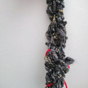 Puede incluir: Un ovillo de hilo texturizado en tonos grises y negros, entrelazado con hilos metálicos dorados. Acentos de hilo rojo están atados alrededor del ovillo. El hilo parece suave y esponjoso, adecuado para tejer u otras manualidades.
