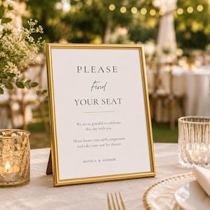 May include: A gold-framed sign with the text "PLEASE Find YOUR SEAT" on a table set for a wedding. The sign is surrounded by a candle, flowers, and glassware. The sign also includes the names "MONICA & ANDREW".