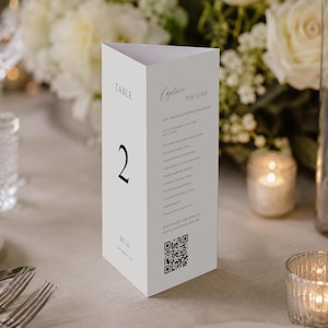 May include: A white, triangular table card with the number 2 in black. The card has text that reads "Capture the Love" and a QR code. The card is on a table with flowers and candles.