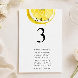 May include: A white table number card featuring a watercolour lemon design and the number 3 in black. Guest names are listed below. The background includes flowers and a wine glass. Measurements are not applicable.