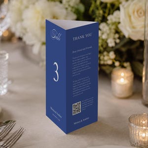 Puede incluir: Una tarjeta con el número de mesa azul marino con el número 3 en blanco. La tarjeta es triangular y presenta las palabras "Table" y "THANK YOU" en blanco. La tarjeta también incluye texto y un código QR. La tarjeta está sobre una mesa con flores y velas.