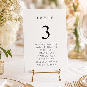 May include: A white table number card with the number 3 in a bold, black font. The card lists names below the number. The card is held in a gold-coloured metal stand. The background includes flowers, candles, and glassware.