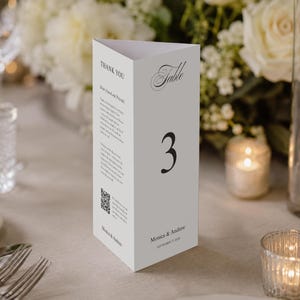 Op de afbeelding: Een witte, driehoekige tafelnummerkaart met het nummer 3 in zwart schrift. De kaart heeft de tekst "Table" en "Thank You". De kaart staat op een tafel met bloemen en kaarsen.