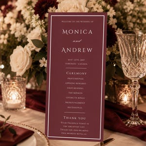 May include: A burgundy wedding ceremony program with white text, featuring the names "Monica and Andrew" and ceremony details. The program is set on a table with floral arrangements, candles, and crystal glassware.