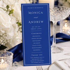 Könnte beinhalten: Ein blaues Hochzeitsprogramm mit weißem Text, mit den Namen "Monica und Andrew" und Zeremoniedetails. Das Programm liegt auf einem Tisch mit weißer Tischdecke, Kristallgläsern und einem Strauß weißer Blumen.