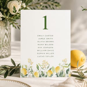 May include: A white table card with a green number 1 at the top and a list of names below. The bottom of the card features a watercolor design of lemons, leaves, and flowers. A lemon and a wine glass are visible in the background.
