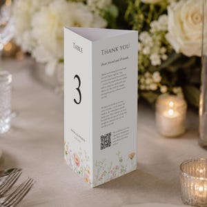 Peut inclure: Un marque-table triangulaire blanc affichant le chiffre 3, le texte "TABLE", "THANK YOU", "MONICA & ANDREW" et une date. La carte comprend un code QR et une illustration florale délicate en bas avec des touches botaniques roses, jaunes et vertes.