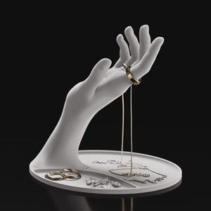 May include: A matte white jewellery display stand featuring a sculpted hand rising from an oval tray. The hand holds a gold ring on one finger, while the tray contains two additional rings, a silver pendant, and a delicate gold chain necklace.