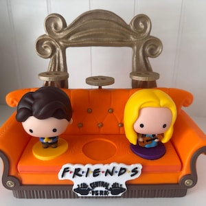 May include: An orange miniature sofa with two cartoon figures, one with brown hair and the other with blonde hair. The sofa has a gold-coloured decorative back and the text "FRIENDS" and "CENTRAL PERK" on the front.