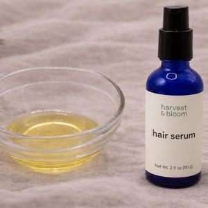 May include: A blue glass bottle with a black pump dispenser containing hair serum, labeled harvest & bloom with a net weight of 2 fl oz (50 g), positioned next to a small clear glass bowl filled with a golden yellow liquid oil.