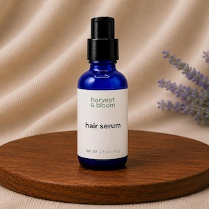 May include: A cobalt blue glass bottle with a black pump dispenser containing hair serum. The white label features the text harvest & bloom, hair serum, and Net Wt. 2 fl oz (50 g) in black lettering. The bottle is cylindrical with a flat base.