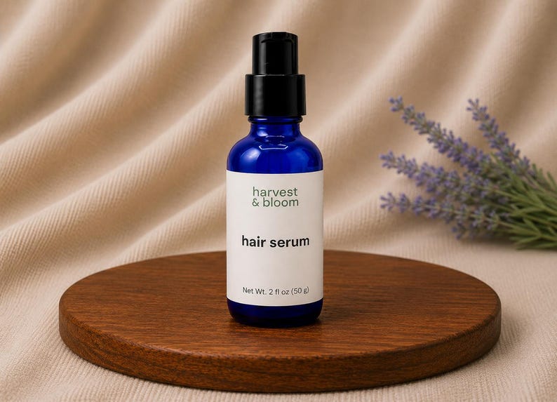 May include: A cobalt blue glass bottle with a black pump dispenser containing hair serum. The white label features the text harvest & bloom, hair serum, and Net Wt. 2 fl oz (50 g) in black lettering. The bottle is cylindrical with a flat base.